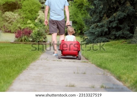 Beautiful young girl sitting in a red wagon cart outdoors. Father is pulling a red wagon with his daughter. Royalty-Free Stock Photo #2313327519