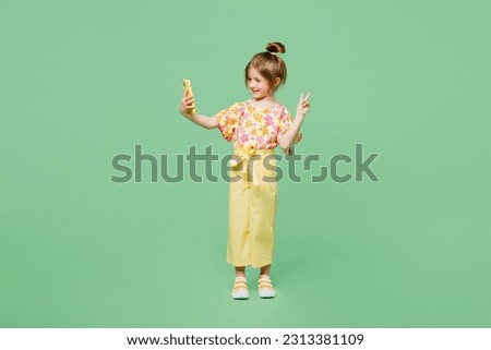 Full body little child kid girl 6-7 years old wear casual clothes doing selfie shot on mobile cell phone e show v-sign isolated on plain green background. Mother's Day love family lifestyle concept