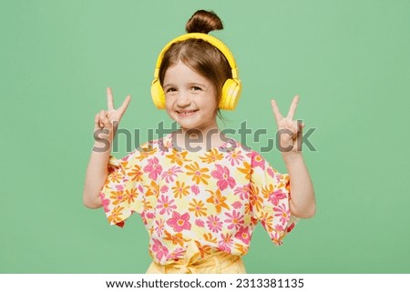 Little fun child kid girl 6-7 year old wears casual clothes headphones listen music show v-sign isolated on plain pastel green background studio portrait. Mother's Day love family lifestyle concept