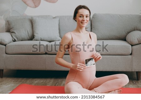 Young sporty fitness trainer instructor pregnant woman wear pink tracksuit hold ultra sound picture train yoga exercises sit stretch on mat floor at home gym indoor. Workout sport health care concept