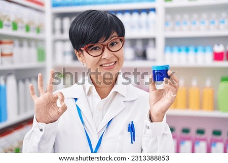 Young asian woman with short hair working at pharmacy drugstore holding cream jar doing ok sign with fingers, smiling friendly gesturing excellent symbol 