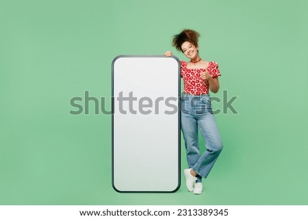 Full body young smiling happy woman wearing casual clothes red blouse big huge blank screen mobile cell phone smartphone with area show thumb up isolated on plain pastel light green color background