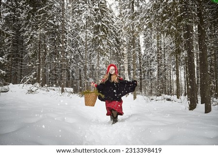Cute little girl in red cap or hat and black coat with basket of green fir branches in forest and snow on winter day. Young girl went to gather firewood in the cold. Fun and fairytale on photo shoot