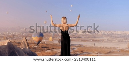 Traveler woman in black dress background landscape Cappadocia with hot air balloons sun light. Concept trip Turkey travel.