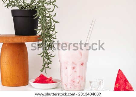 Glass of tasty refreshing cold summer drink with watermelon, watermelon slice, ice cube, straw and plant on white background with copy space. Layout for wide banner.