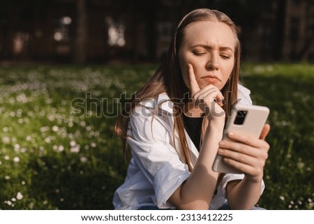 Woman holding a phone while holding her hand on chin. Expressive student thinking outside in campus - Woman in doubt with smartphone in her palm, think about reading information, search solution. Royalty-Free Stock Photo #2313412225