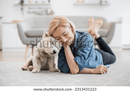 Kind female person and faithful small terrier snuggling to each other with heads while staying on wooden floor at home. Affectionate woman and cuddly Westie enjoying bonding interaction on sunny day. Royalty-Free Stock Photo #2313431223