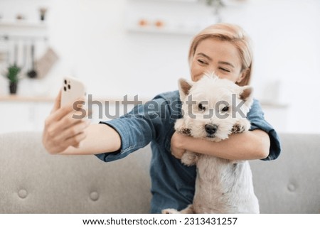 Close up view of vivid blonde lady in denim shirt looking at front camera of mobile while hugging smart terrier indoors. Lovely woman getting self-taken picture with furry friend on room background.