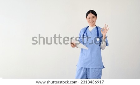 A professional and beautiful young Asian female doctor or medical worker in scrubs showing the Okay hand sign and holding a medical clipboard. isolated on a white background. Good job, Awesome