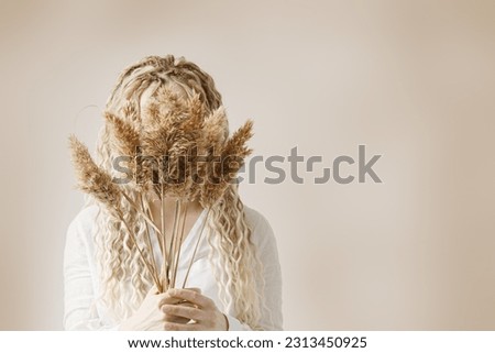 Woman holding pampas grass in front of face, light background, autumn, fall minimal trend concept. Female with long hair dreadlocks hiding behind bouquet dried flowers. Monochrome lifestyle photo