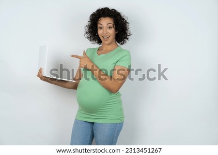Shocked Beautiful pregnant woman wearing green T-shirt standing over white studio background pointing finger modern device