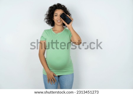 Beautiful pregnant woman wearing green T-shirt standing over white studio background holding modern smartphone covering one eye while smiling