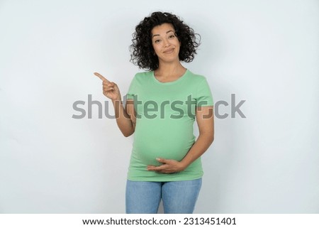 Beautiful pregnant woman wearing green T-shirt standing over white studio background points to side on blank space demonstrates advertisement. People and promotion concept