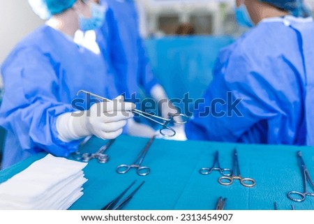 Nurse hand taking surgical instrument for group of surgeons at background operating patient in surgical theatre. Steel medical instruments ready to be used. Surgery and emergency concept Royalty-Free Stock Photo #2313454907