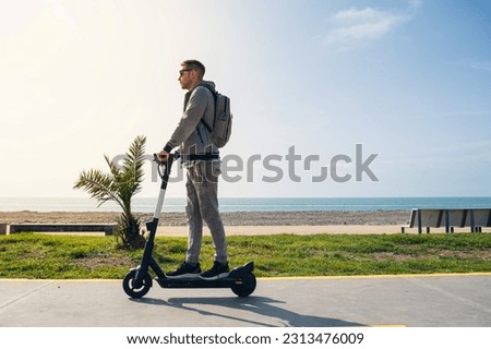 Young man traveler riding e-scooter along the sea coast and beach at sunny summer day. Male driving electric scooter outdoors. Sustainable lifestyle concept
