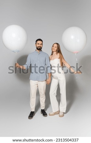 Full length of fashionable and happy expecting parents holding white balloons and looking at camera during gender reveal surprise party on grey background