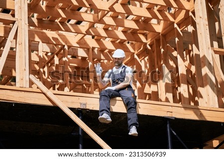 Man architect on construction site of wooden-framed house gestures the OK sign. The concept pertains to modern ecological construction.