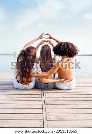 Three female teenagers of different ethnic groups show heart symbols with their hands. Teen girls in casual clothes sit on wooden bridge near the river or lake and hug. Friendship concept. Copy space