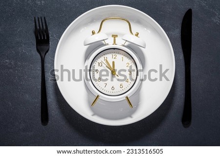 White plate and clock on a dark background with copy space. Concept of interval fasting and autophagy. Royalty-Free Stock Photo #2313516505