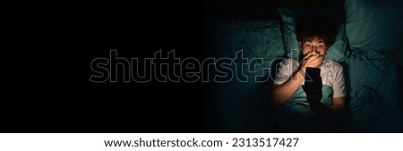 Young scared guy lying on bed and watching horror movie on mobile phone in dark bedroom in late evening. Banner, copy space Royalty-Free Stock Photo #2313517427