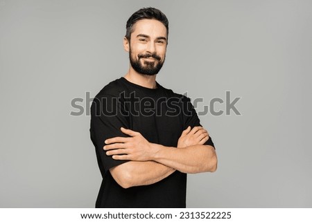 Smiling brunette and bearded man in casual black t-shirt crossing arms and looking at camera while standing isolated on grey, masculine beauty concept, confident and charismatic Royalty-Free Stock Photo #2313522225
