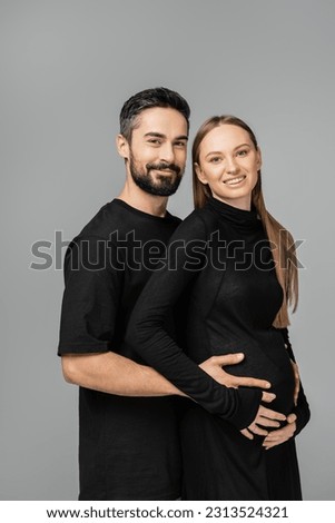 Portrait of bearded man in black t-shirt looking at camera while touching belly of smiling and stylish pregnant wife in dress isolated on grey, growing new life concept