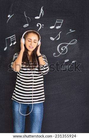 A shot of attractive young woman standing in front of a blackboard with musical notes drawn on and listening to music on headphones