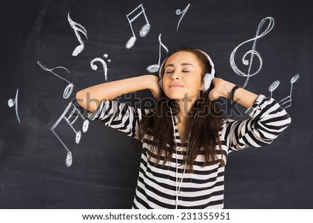 A shot of attractive young woman standing in front of a blackboard with musical notes drawn on and listening to music on headphones