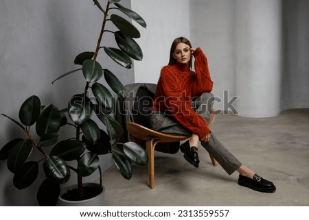 High fashion photo of beautiful elegant young woman in pretty orange terracotta sweater, gray pants, trousers posing on textured wall, tall flower. Slim figure. Model is sitting on a chair.