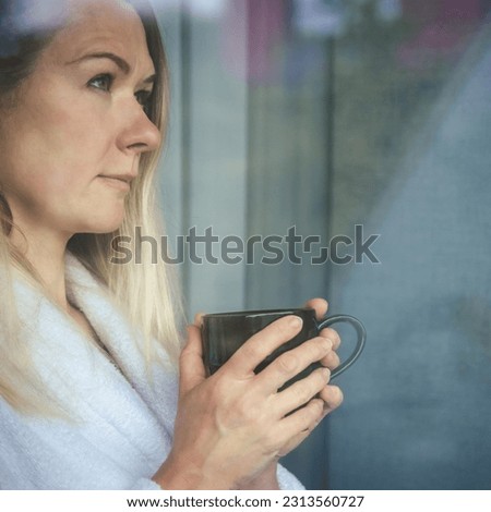 Woman in elegant robe drinking coffee in hotel room and standing near window. face closeup, window reflection