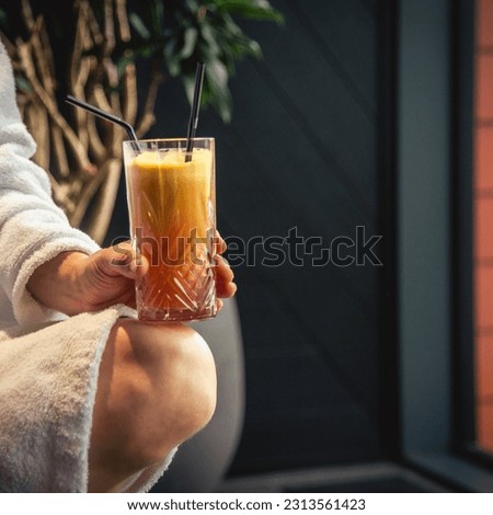 Woman in white bathrobe lying on sofa and relaxing with orange cocktail at home near window