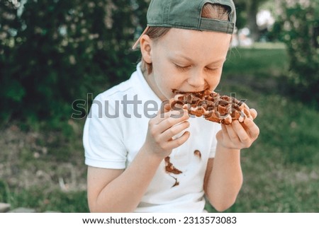  A boy eating a sweet pastry covered with chocolate. dirty stain on clothes. outdoors. High quality photo