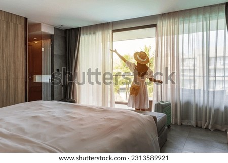 Young woman traveler opening the curtains and looking at the view from the window of a hotel room while on summer vacation, Travel lifestyle concept Royalty-Free Stock Photo #2313589921
