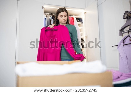 Woman chooses clothes for a celebration, holds a pink birthday dress in her hands. Cluttering the wardrobe, charity, clothes box, organization and storage of things, apartment, relocation