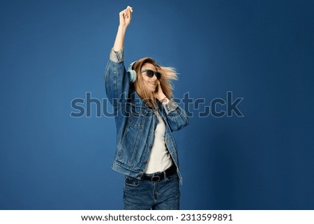 Portrait of young girl in jeans outfit listening to music in headphones, smiling, dancing against blue studio background. Concept of youth, human emotions, facial expression, fun, leisure