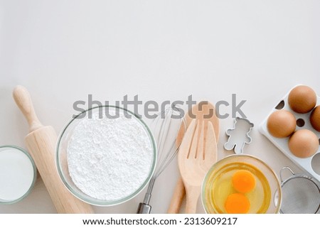 Close-up top view of fresh baking ingredients, eggs, and flour and kitchen wares, utensils on white background table for preparation of making delicious pastry.