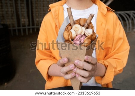 Hong Kong or bubble waffle with ice cream, banana and chocolate tubes in the hands of a child Royalty-Free Stock Photo #2313619653