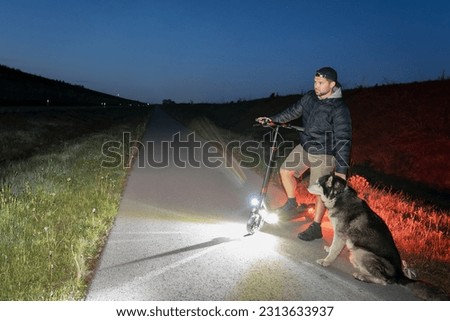 A man rides an electric scooter at night with a husky dog, low light photo.