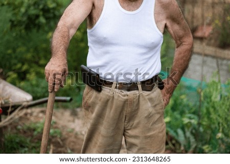 unrecognisable man in the orchard, a retired man in his eighties with a tank top leaning on a hoe, corduroy trousers and a mobile phone in a leather holster on his belt. Royalty-Free Stock Photo #2313648265