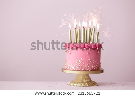 Pink birthday cake with gold birthday candles and celebration sparklers against a plain pink background Royalty-Free Stock Photo #2313663721