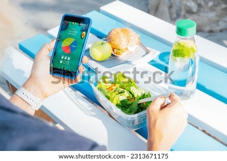 Close up woman using meal tracker app on phone while eating salad at picnic table in the park on a break. Healthy balanced diet lunch box. Healthy diet plan for weight loss. Selective focus Royalty-Free Stock Photo #2313671175