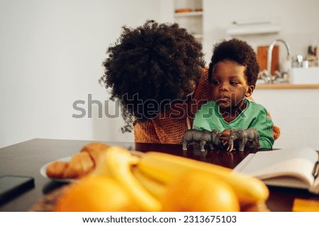 An interracial boy is sitting at the breakfast table in his mother's lap and has a serious face. There are toys and breakfast on the table. A little boy is sulking. Diverse family at home.