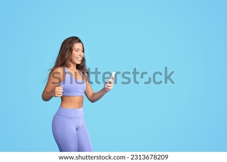 Glad millennial caucasian slim woman in sportswear with smartphone has fun, isolated on blue studio background. Sports app, blog for weight loss and body care, good news and jump