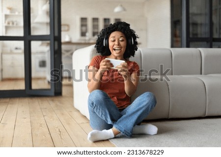Excited Black Woman Playing Video Games On Smartphone At Home, Happy Cheerful African American Female Holding Mobile Phone And Celebrating Online Win While Sitting On Floor In Living Room, Copy Space