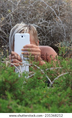 A girl shooting with smart phone hiding in the bushes outdoors