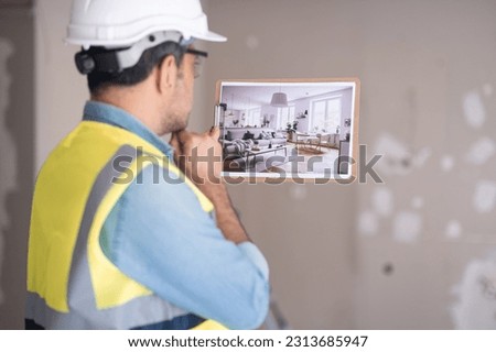 Pensive engineer in protective helmet thinking about Scandinavian interior design while looking at picture in hand against shabby wall in living room renovation process in apartment