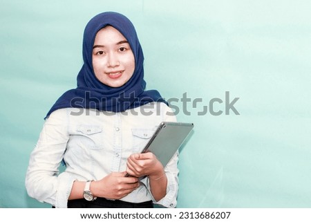 Portrait beautiful young Asian Muslim woman wearing hijab and casual outfit holding digital tablet and pointing finger at copy space isolated on blue studio background
