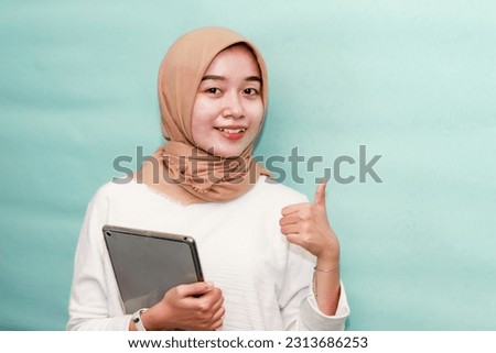 Portrait beautiful young Asian Muslim woman wearing hijab and casual outfit holding digital tablet and pointing finger at copy space isolated on blue studio background