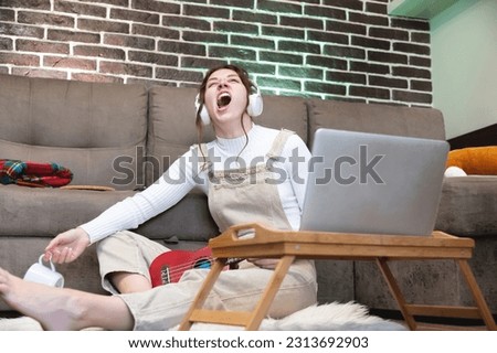 young woman yawning after a long working day sitting in front of a laptop wearing wireless headphones with a cup in her hand in an apartment