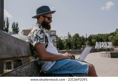 Photo of a freelance web designer young guy working outdoors on a laptop computer connected to a public Wi-Fi. Carefree caucasian hipster spends his free time in the park.